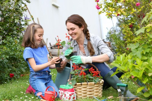 A woman and young girl gardening together in a lush outdoor space, surrounded by green plants, shrubs, and flowering bushes. The woman, dressed casually in a light grey shirt and green gardening gloves, is smiling as she hands a small potted plant with red flowers to the girl, who is wearing a blue t-shirt and denim shorts. They are seated on the grass near a variety of garden tools and a woven basket, with a watering can and decorated fabric container nearby. The garden features a neatly maintained lawn, a mix of flowering plants, and dense foliage, indicative of a well-kept residential garden in Shadwell, with bright natural light suggesting a clear day. This scene reflects outdoor gardening activities typical of local gardening services, emphasizing nurturing plants and garden maintenance in a welcoming outdoor environment.