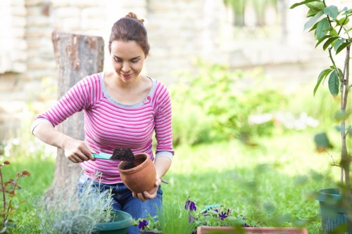 A young woman in a pink and white striped long-sleeve top is engaged in gardening in a landscaped garden with lush greenery and a stone wall in the background. She is holding a small terracotta pot filled with soil while using a trowel to perform planting or soil management. In front of her, there are various potted plants, including flowering species with purple blooms, and a garden bed with plants and shrubs. The garden features a well-maintained grassy lawn with a mixture of flowering and foliage plants, bordered by a paved pathway or decking area partially visible at the edge of the image. The scene is lit with natural daylight, suggesting a mild, cloudy outdoor environment typical of spring or summer, and reflects professional gardening and landscaping services offered by Lawn Mowing Shadwell in the Shadwell area, with the garden designed for both aesthetic appeal and outdoor enjoyment.
